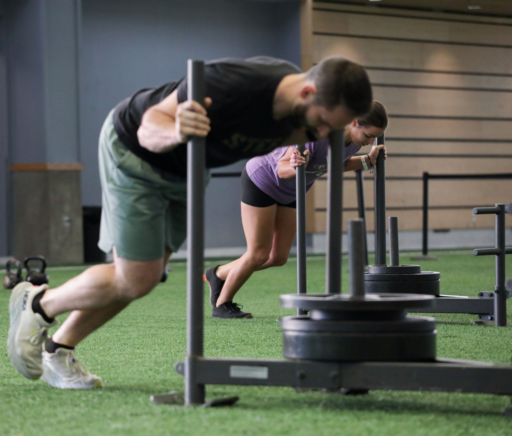 075A5099 A man and a woman push weighted sleds across green indoor turf in a gym, leaning forward with both hands gripping the vertical poles as they perform a strength and conditioning workout. Kettlebells and gym equipment are visible in the background.