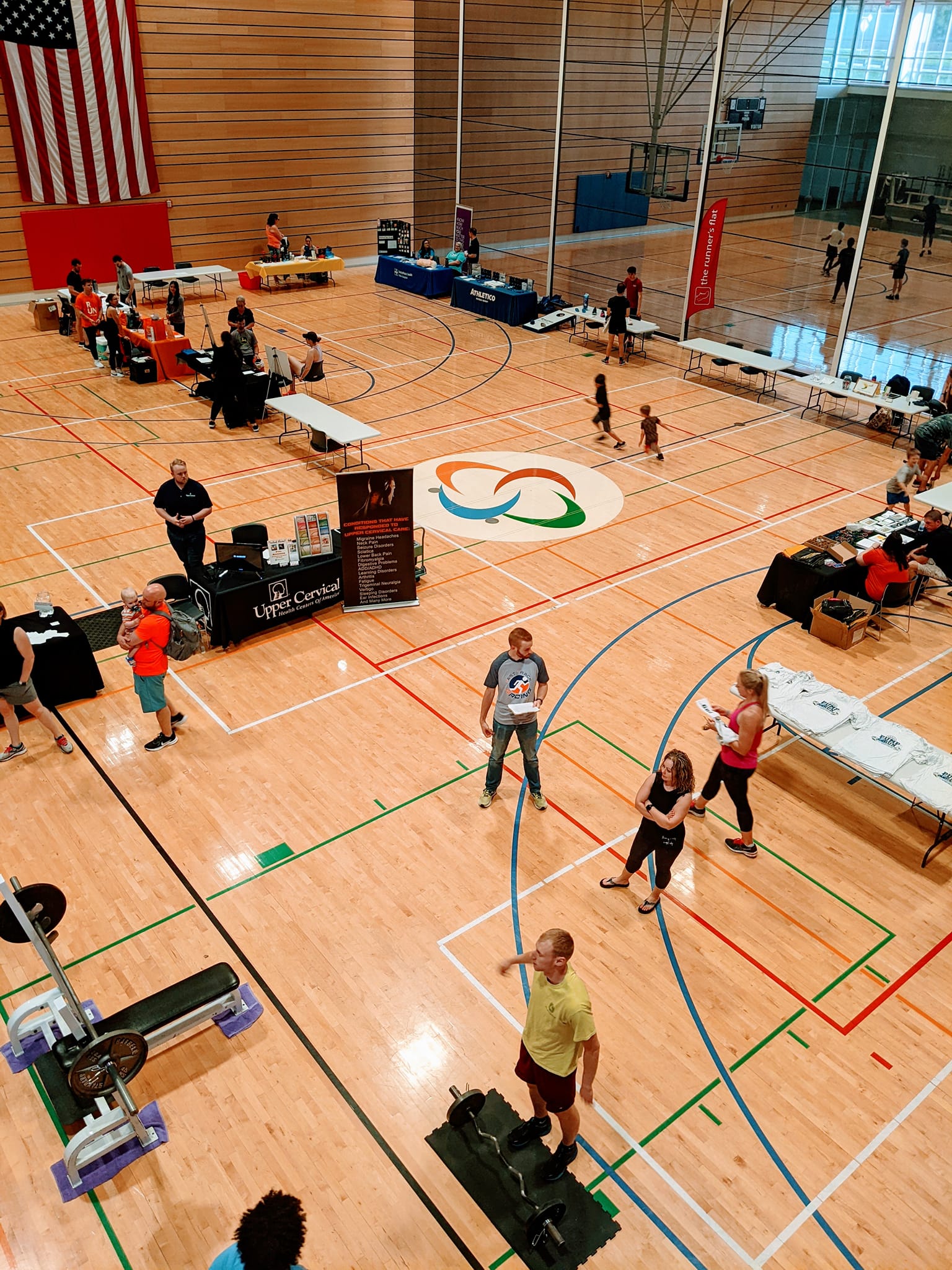health expo photo people standing around a variety of vendor tables that are set up on a basketball court during a health expo.
